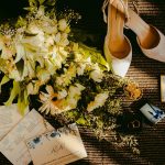 Wedding flat lay featuring shoes, flowers, and invitations with a golden tone.